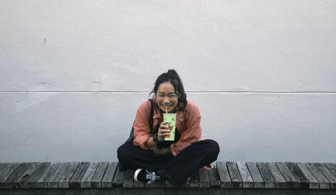 Maggie Chang smiling on a wooden bench against a white wall, holding a green drink cup.
