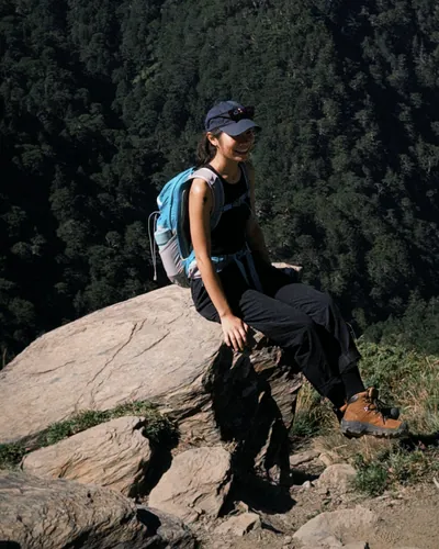 Maggie sitting on a rock above a forested mountain valley, wearing a cap and a blue backpack.