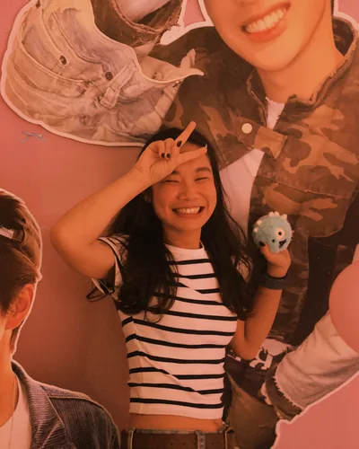 Maggie grinning and making a peace sign next to life-size K-pop idol cutouts at a pink fan café.