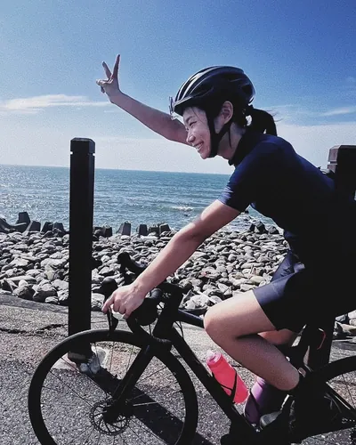 Maggie cycling along a seaside road, waving a peace sign with the ocean behind her.